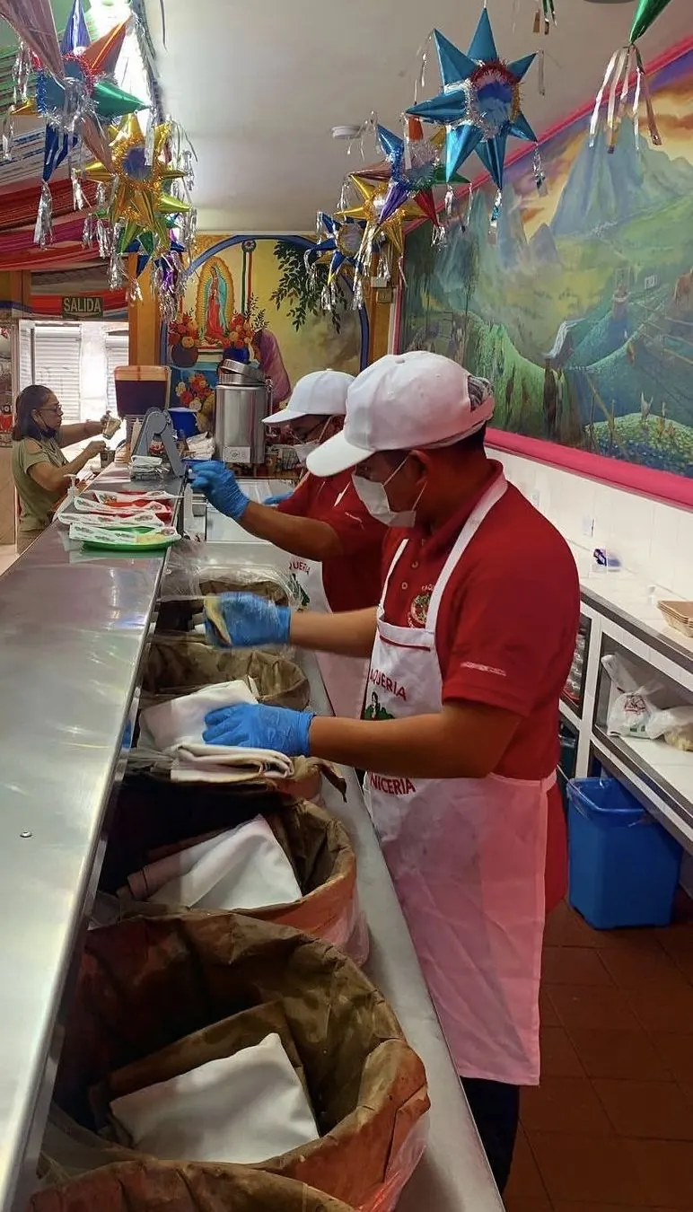 Taquero sirviendo tacos de canasta en la barra de La Mexicana, con el mural detrás